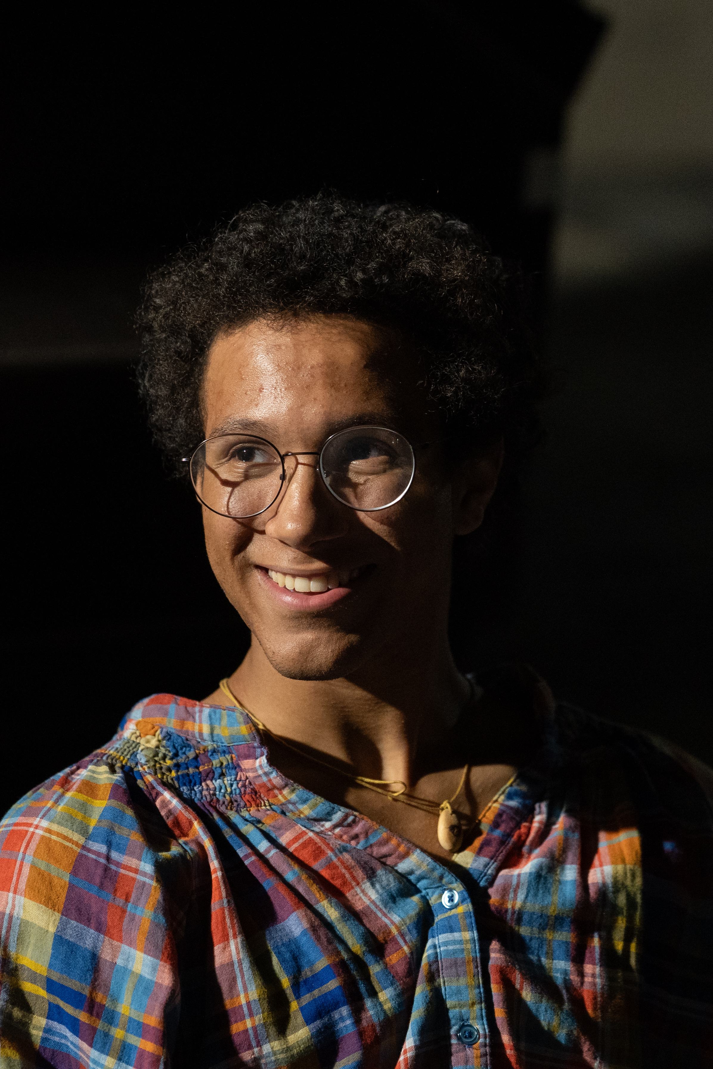 Portrait of a smiling young man with curly hair and round glasses, in a plaid shirt, lit from the side