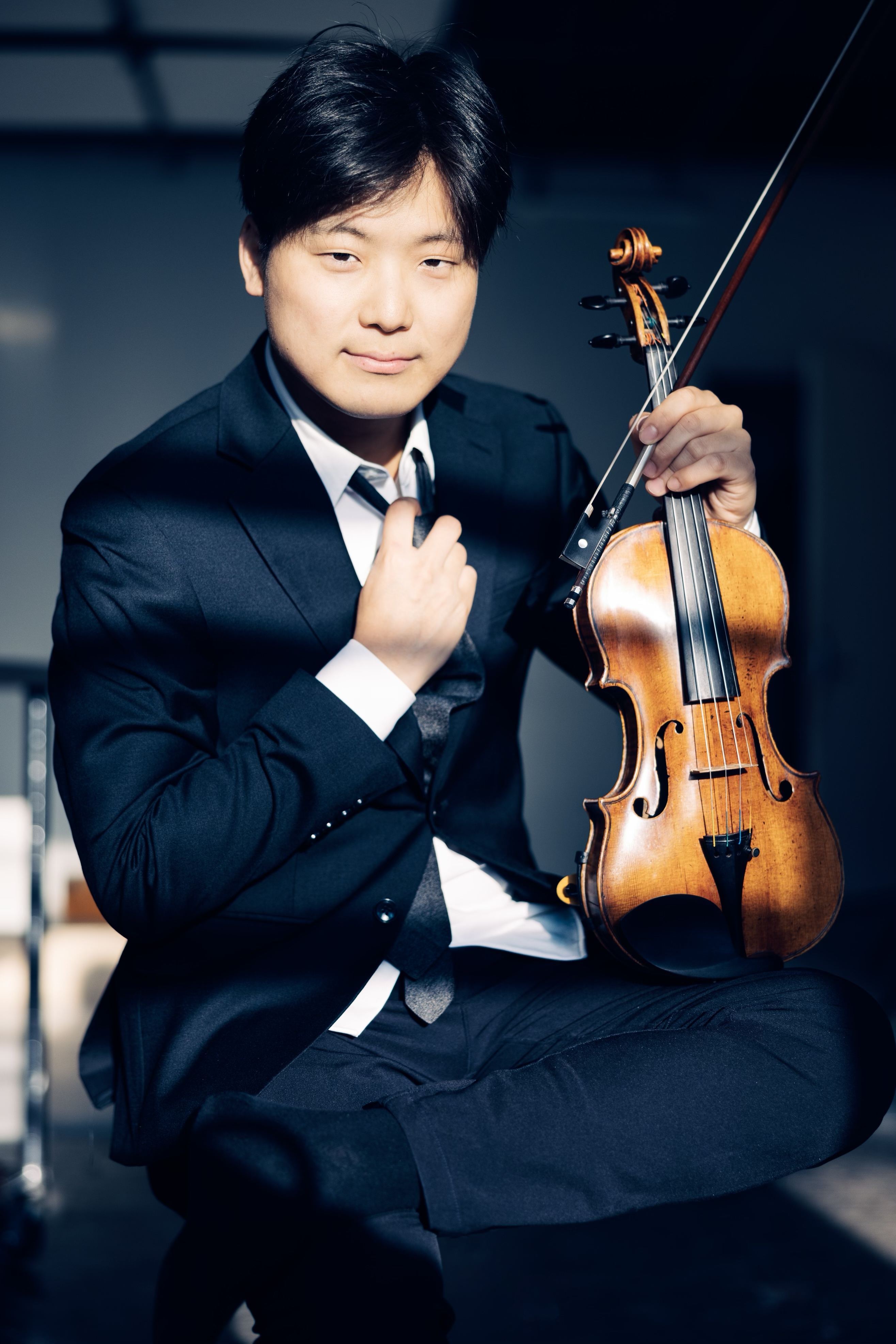 Siwoo Kim in a dark suit in window light, holding his violin and bow