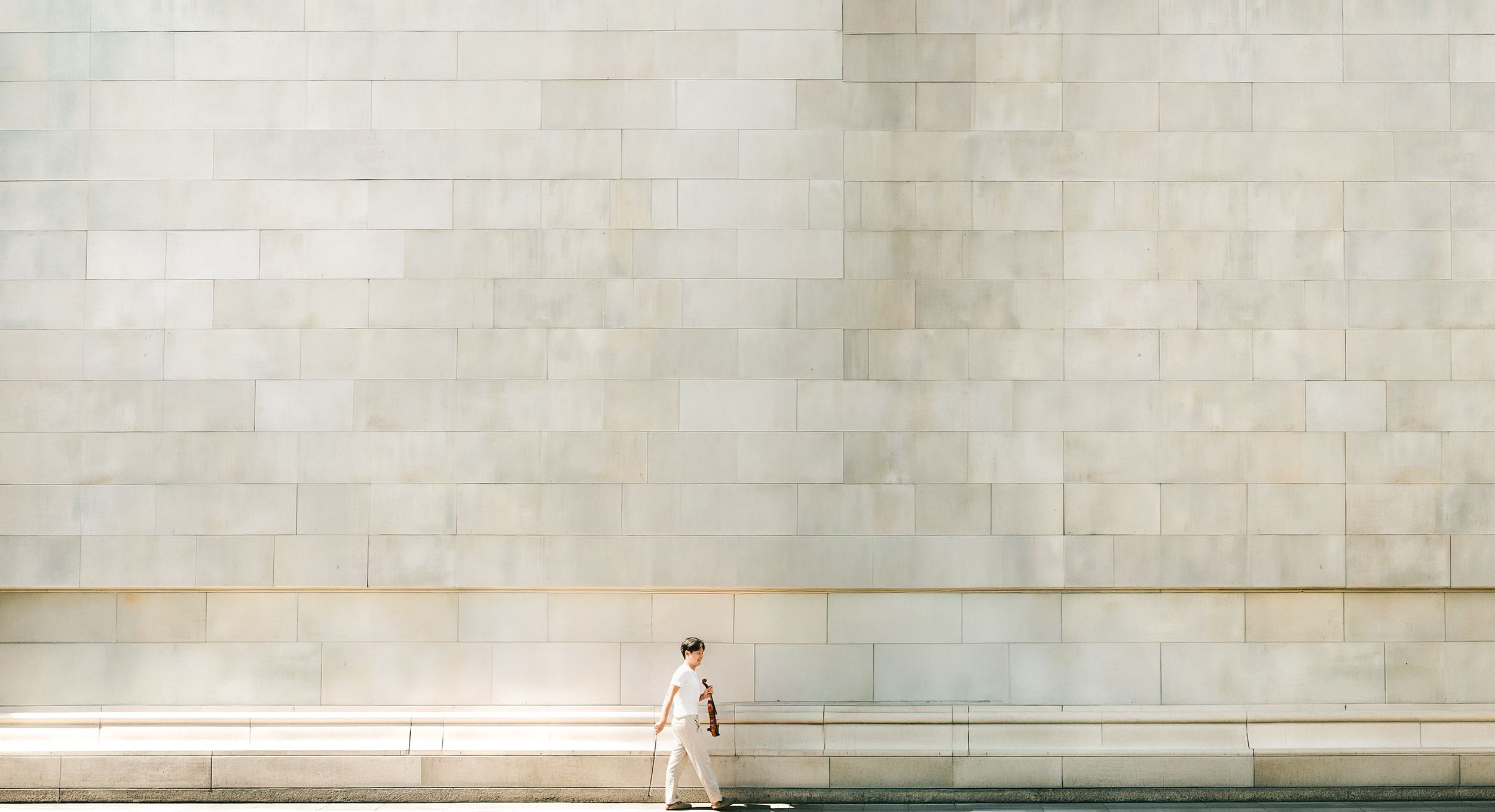 Siwoo Kim walking past a long limestone wall, violin and bow in hand